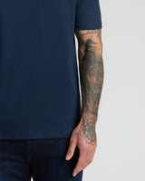 Close-up of a man's arm with detailed tattoos wearing a Navy Pima Cotton Blend Crew shirt paired with dark jeans against a plain light background.